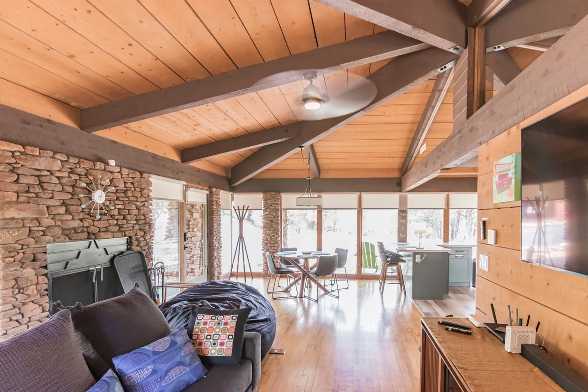 A comfortable living space featuring a stone fireplace, dining area, and expansive windows that frame the natural world outside. Wooden beams and a bean bag chair create a relaxed, rustic atmosphere, inviting guests to enjoy both cozy winter evenings and sunny afternoons with views of the landscape.