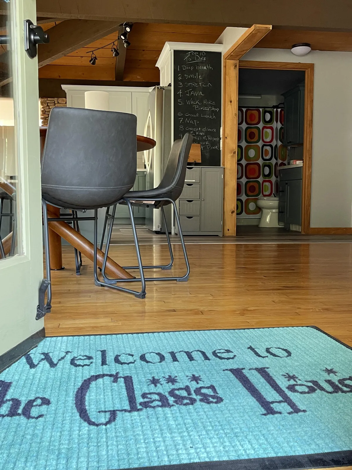 A welcoming entrance scene with a visible welcome mat, chalkboard, dining chairs, and a glimpse of the open kitchen area, evoking the feeling of returning home after a day out exploring local shops and eateries.