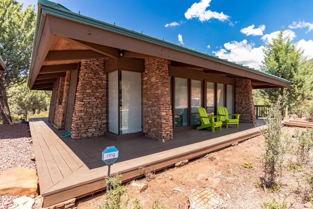This image features an inviting wooden deck furnished with green chairs and stone columns, set against the tranquil backdrop of surrounding trees. The view from the deck offers a seamless transition from interior comfort to the serenity of the outdoors, suggesting the perfect spot to observe wildlife or enjoy a peaceful morning coffee while soaking in the natural environment.