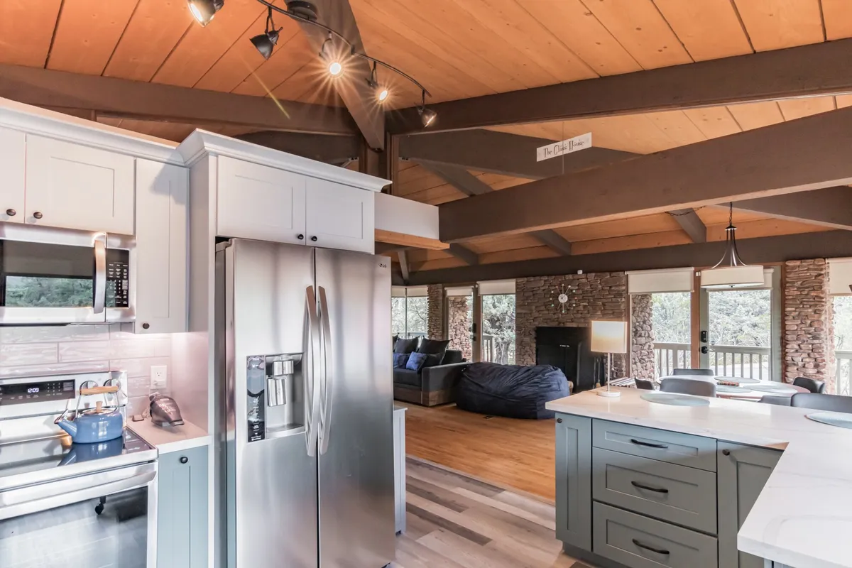A view of a spacious, modern kitchen featuring sleek appliances, an open floor plan, a cozy stone fireplace, exposed wooden beams, and large windows that flood the space with natural light. The kitchen radiates warmth, ideal for gathering and cooking together.