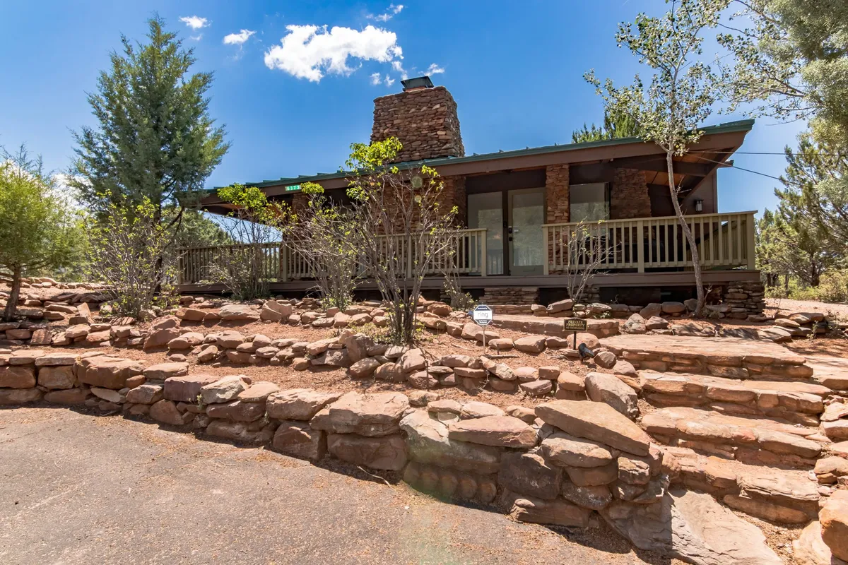 The image depicts a welcoming wooden deck with a sturdy stone chimney, surrounded by rock landscaping and nestled among trees. The porch area is perfectly positioned for enjoying outdoor relaxation and taking in views of the sky as the sun sets over the landscape.