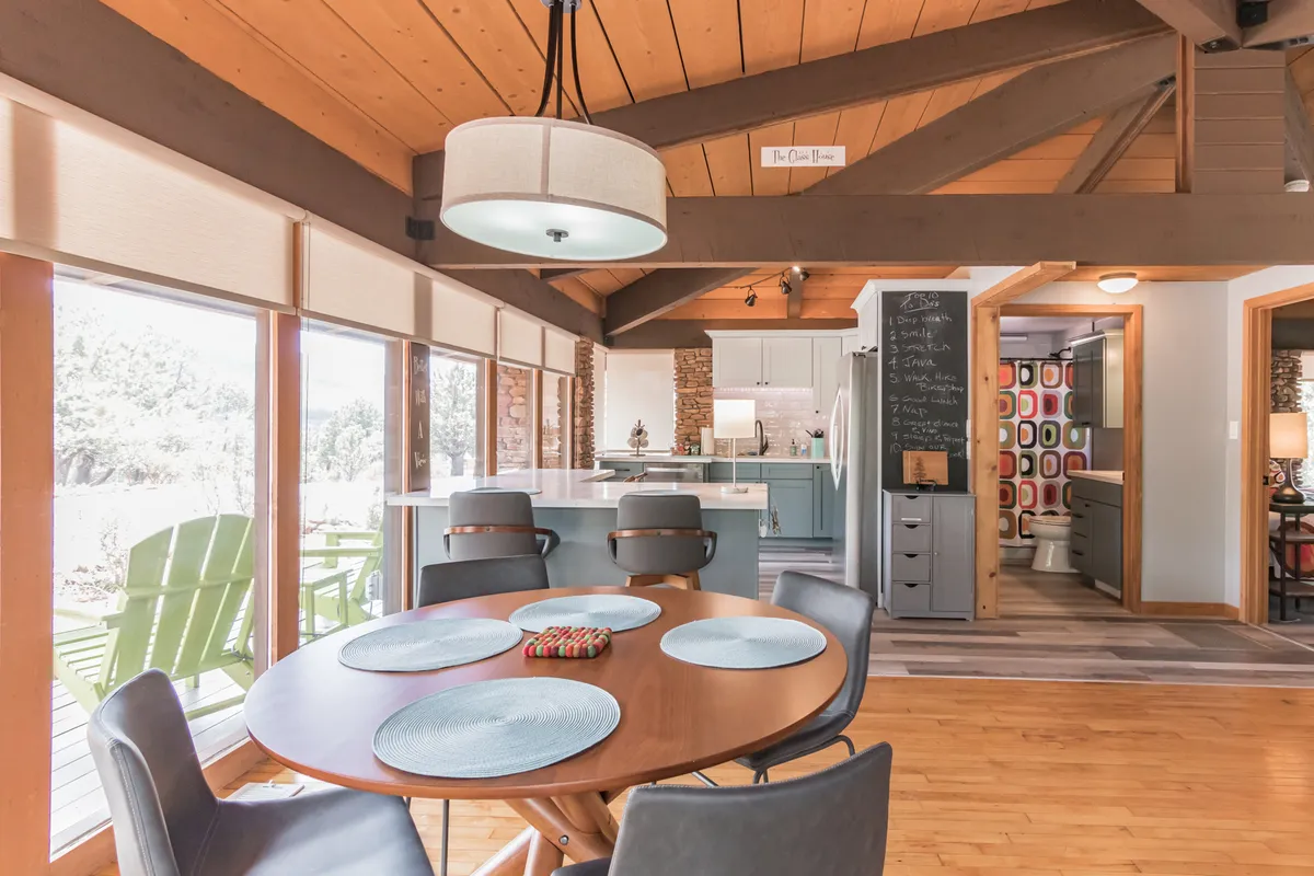 A welcoming dining area with a round table set near large windows, offering views of the landscape beyond. The kitchen is visible in the background, complemented by colorful wall decor that adds vibrancy to the space.