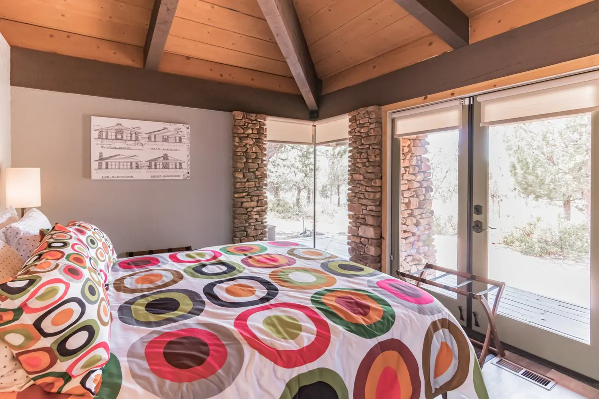 A vibrant bedroom featuring a king-sized bed dressed in colorful bedding with circular patterns, set against a striking stone wall. Large windows flood the space with natural light and offer serene views, creating an inviting atmosphere for relaxation and restful sleep.
