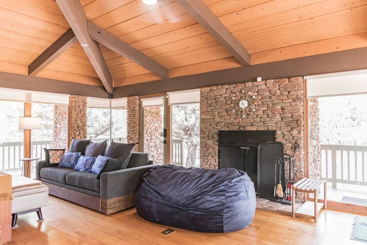 This image showcases a comfortable living area featuring a plush sofa with cushions, an oversized bean bag chair, a stone fireplace, and expansive floor-to-ceiling windows that flood the space with natural light and offer unobstructed views of the surrounding landscape.
