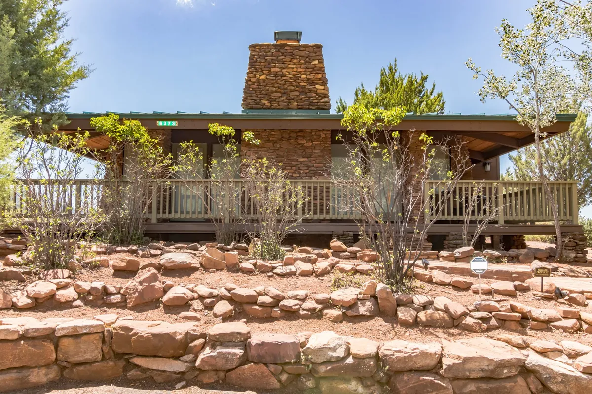 This image shows a tranquil exterior view featuring a wooden wrap-around deck attached to the house, with a prominent stone chimney, landscaped gardens, and lush greenery all around. The deck looks out onto the natural surroundings, ideal for relaxing and enjoying scenic views.