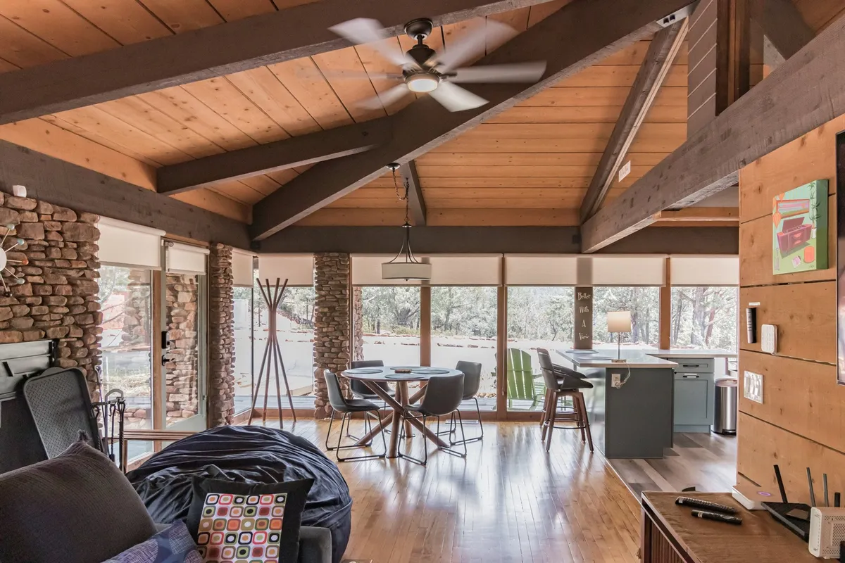 A cozy dining area with exposed wooden beams, stone walls, and large windows overlooking the surrounding forest. The table is set for casual gatherings, and colorful throw pillows add warmth. Natural light floods the room, emphasizing the connection between the indoor living space and the forested mountain setting.