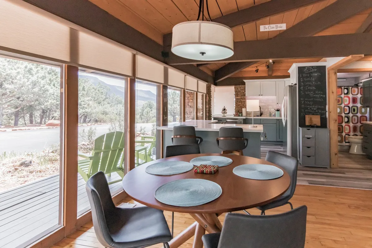 A bright, stylish interior dining area with a table set by large windows, filling the space with natural light and offering glimpses of the outdoor scenery. The decor is contemporary and inviting, hinting at intimate dinners and cozy indoor evenings.